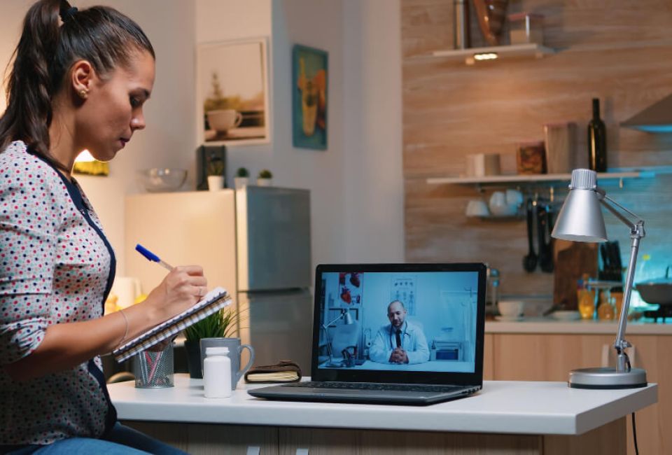 A woman sits at a desk, focused on her laptop, with a notepad and pen beside her.