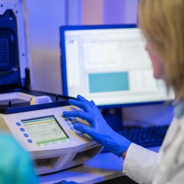 A laboratory technician wearing blue gloves operates a thermal cycler beside a computer displaying test data.