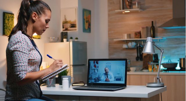 A woman sits at a desk, focused on her laptop, with a notepad and pen beside her.