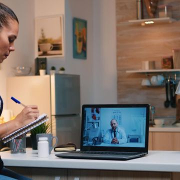 A woman sits at a desk, focused on her laptop, with a notepad and pen beside her.